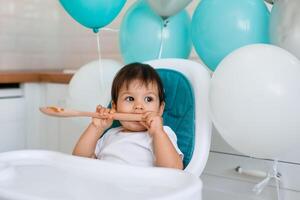 Little baby boy sitting in blue high chair at home on white kitchen and playing with wooden big spoon on background with balloons. photo