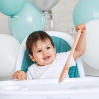 Little baby boy sitting in blue high chair at home on white kitchen and playing with wooden big spoon on background with balloons. photo