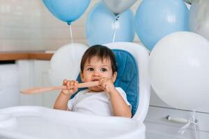 Little baby boy sitting in blue high chair at home on white kitchen and playing with wooden big spoon on background with balloons. photo
