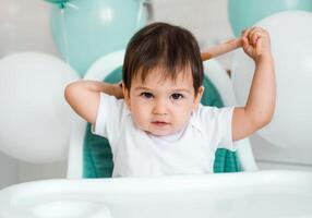 Little baby boy sitting in blue high chair at home on white kitchen and playing with wooden big spoon on background with balloons. photo
