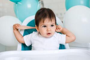 Little baby boy sitting in blue high chair at home on white kitchen and playing with wooden big spoon on background with balloons. photo