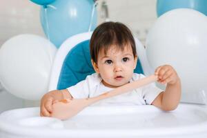 Little baby boy sitting in blue high chair at home on white kitchen and playing with wooden big spoon on background with balloons. photo