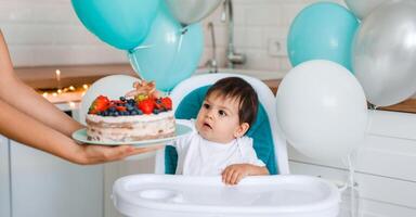 Little baby boy sitting in high chair in white kitchen and tasting first year cake with fruits from mother hands on background with balloons. photo