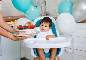 Little baby boy sitting in high chair in white kitchen and tasting first year cake with fruits on background with balloons. photo