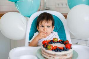 Little baby boy sitting in high chair in white kitchen and tasting first year cake with fruits on background with balloons. photo