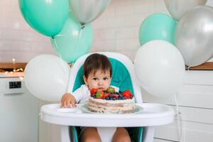 Little baby boy sitting in high chair in white kitchen and tasting first year cake with fruits on background with balloons. photo