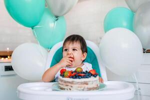 Little baby boy sitting in high chair in white kitchen and tasting first year cake with fruits on background with balloons. photo