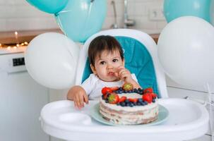 Little baby boy sitting in high chair in white kitchen and tasting first year cake with fruits on background with balloons. photo