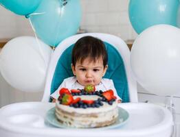 Little baby boy sitting in high chair in white kitchen and tasting first year cake with fruits on background with balloons. photo
