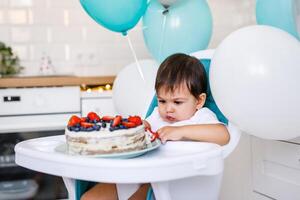 Little baby boy sitting in high chair in white kitchen and tasting first year cake with fruits on background with balloons. photo