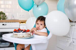 Little baby boy sitting in high chair in white kitchen and tasting first year cake with fruits on background with balloons. photo