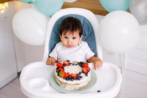 Little baby boy sitting in high chair in white kitchen and tasting first year cake with fruits on background with balloons. photo