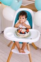 Little baby boy sitting in high chair in white kitchen and tasting first year cake with fruits on background with balloons. photo