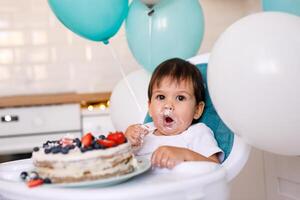 Little baby boy sitting in high chair in white kitchen and tasting first year cake with fruits on background with balloons. photo