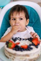 Little baby boy sitting in high chair in white kitchen and tasting first year cake with fruits on background with balloons. photo
