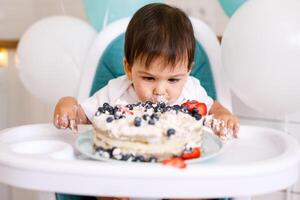 Little baby boy sitting in high chair in white kitchen and tasting first year cake with fruits on background with balloons. photo