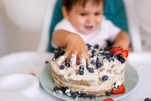 Little baby boy sitting in high chair in white kitchen and tasting first year cake with fruits on background with balloons. photo