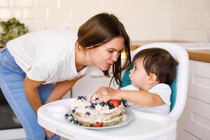 Happy mother with one year old baby celebrating with first birthday with white kitchen on background photo