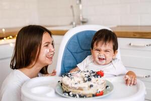 Happy mother with one year old baby celebrating with first birthday with white kitchen on background photo