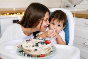 Happy mother with one year old baby celebrating with first birthday with white kitchen on background photo