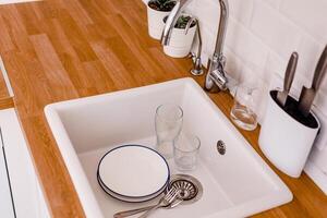 White plates, glass glasses and forks in a clean white sink on a wooden table in a white kitchen photo