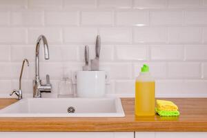 White plates, glass cups and forks in a clean white sink and two bottles of detergent and microfiber rags on a wooden table in a white kitchen photo