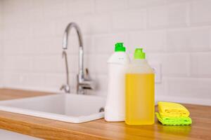 White plates, glass cups and forks in a clean white sink and two bottles of detergent and microfiber rags on a wooden table in a white kitchen photo