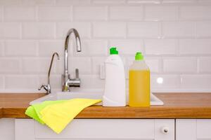 White plates, glass cups and forks in a clean white sink and two bottles of detergent and microfiber rags on a wooden table in a white kitchen photo
