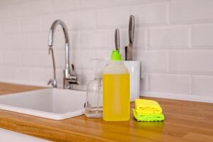 White plates, glass cups and forks in a clean white sink and two bottles of detergent and microfiber rags on a wooden table in a white kitchen photo