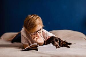 Child wearing glasses reading a book in bed with his dog photo