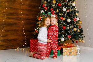 Little brother and sister in red pajamas are standing next to a Christmas tree photo