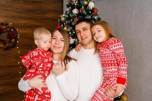Mom, Dad, and the brother and sister in red pajamas are playing and hugging in front of the Christmas tree photo