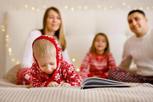 The little boy in festive pajamas is enjoying a book, while his parents and sister can be seen behind him. photo