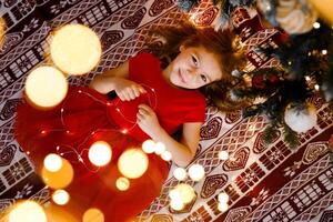 A happy girl in a red dress is lying under the Christmas tree holding a garland, seen from above photo