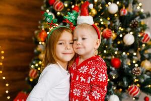Young boy and girl in crimson and white nightwear with playful holiday headbands are standing beside the Christmas tree. photo