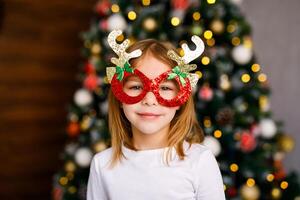 A six-year-old girl in shiny red glasses with golden horns is posing against the background of a Christmas tree photo