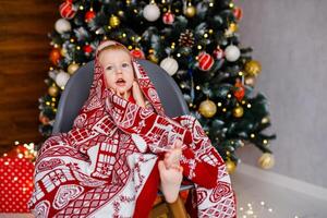 A 2-year-old boy is sitting in a chair wrapped in a warm blanket against the backdrop of a large Christmas tree photo