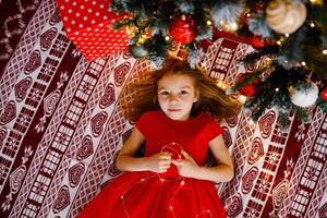 A happy girl in a red dress is lying under the Christmas tree holding a garland, seen from above photo