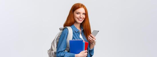 Charismatic friendly-looking european redhead female university student, college girl with backpack and notebooks, heading home after classes, texting friend on smartphone, smile camera photo