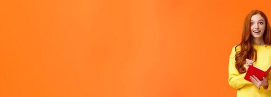Inspired and upbeat, dreamy fascinated redhead girl writing down interesting lecture, smiling and gazing with admiration or enthusiasm, fill-in notebook schedule, standing orange background photo