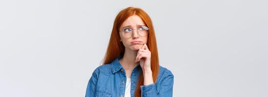 Uneasy sulky cute timid redhead college girl in glasses, look up thoughtful and dreamy, pouting unhappy feeling sad, regret comming party, standing white background indecisive and upset photo