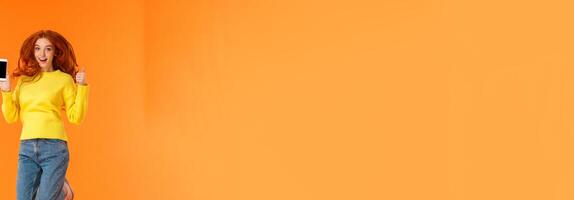 Advertising, technology and holidays concept. Vertical full-length shot cheerful gorgeous ginger girl, redhead woman jumping over orange background with smartphone, show thumb-up photo