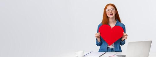Valentines day, creativity and feelings concept. Cheerful smiling redhead girl with long-distance relatioship sending her love through internet, using web cam to show big red heart and say love you photo