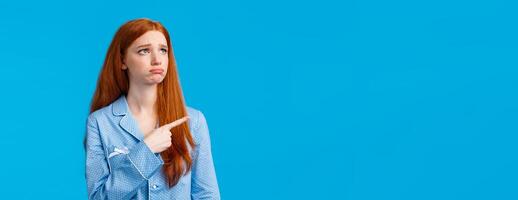 Envy and sad sulking moody redhead teenage girl in nightwear, looking and pointing upper left corner unsatisfied, jealous expression, seeing something she wants, feeling uneasy, blue background photo