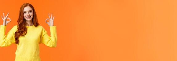 Perfect idea concept, acception. Good-looking redhead girl in yellow sweater, showing okay, good or approval sign smiling nod agreement, satisfied with result, all perfect, orange background photo
