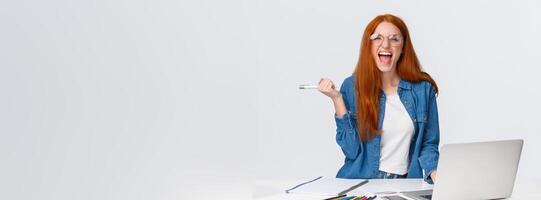 Excited and triumphing good-looking redhead female student passed challenge, found out final exam rating online using laptop, holding pen and screaming happily, standing white background photo