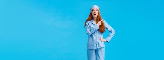 Full-length vertical shot excited and intrigued cheerful redhead female in nightwear and sleep mask, open mouth saying wow, have you seen it, pointing upper left corner amazed, blue background photo