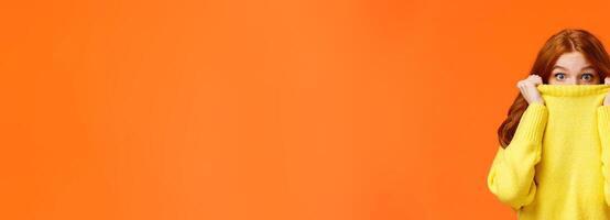 Surprised and astonished cute curious redhead female pulling sweater collar on face and popping eyes at camera with amazed expression, found something interesting, orange background photo