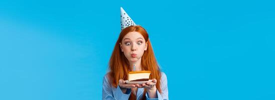 Silly and cute glamour redhead caucasian female in b-day cap and nightwear, holding slice cake with candle, blowing out and making wish with funny expression, standing blue background photo