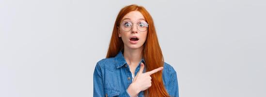 Surprised and wondered, amused attractive redhead woman in glasses, red natural hair, freckles, blue eyes open mouth fascinated and curious, pointing finger left, asking question about banner photo